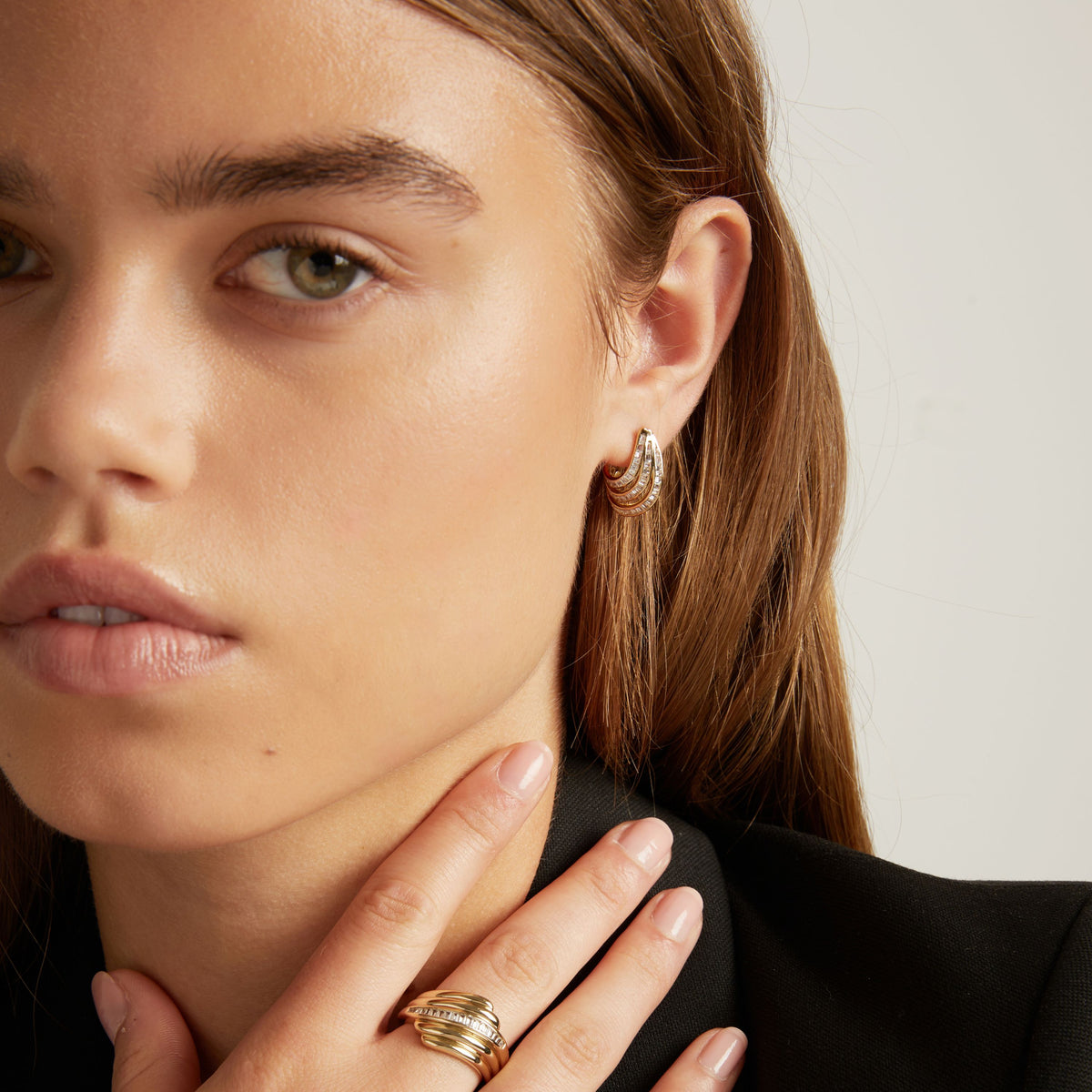 A close-up of a woman with light brown hair wearing a small gold hoop earring with textured, baguette gemstone detailing, set against a plain light gray background.