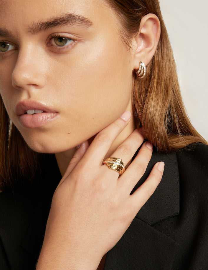 Close-up of a woman wearing a gold ring with textured horizontal bands and a baguette diamonds. Paired with gold hoop earring, a black blazer.