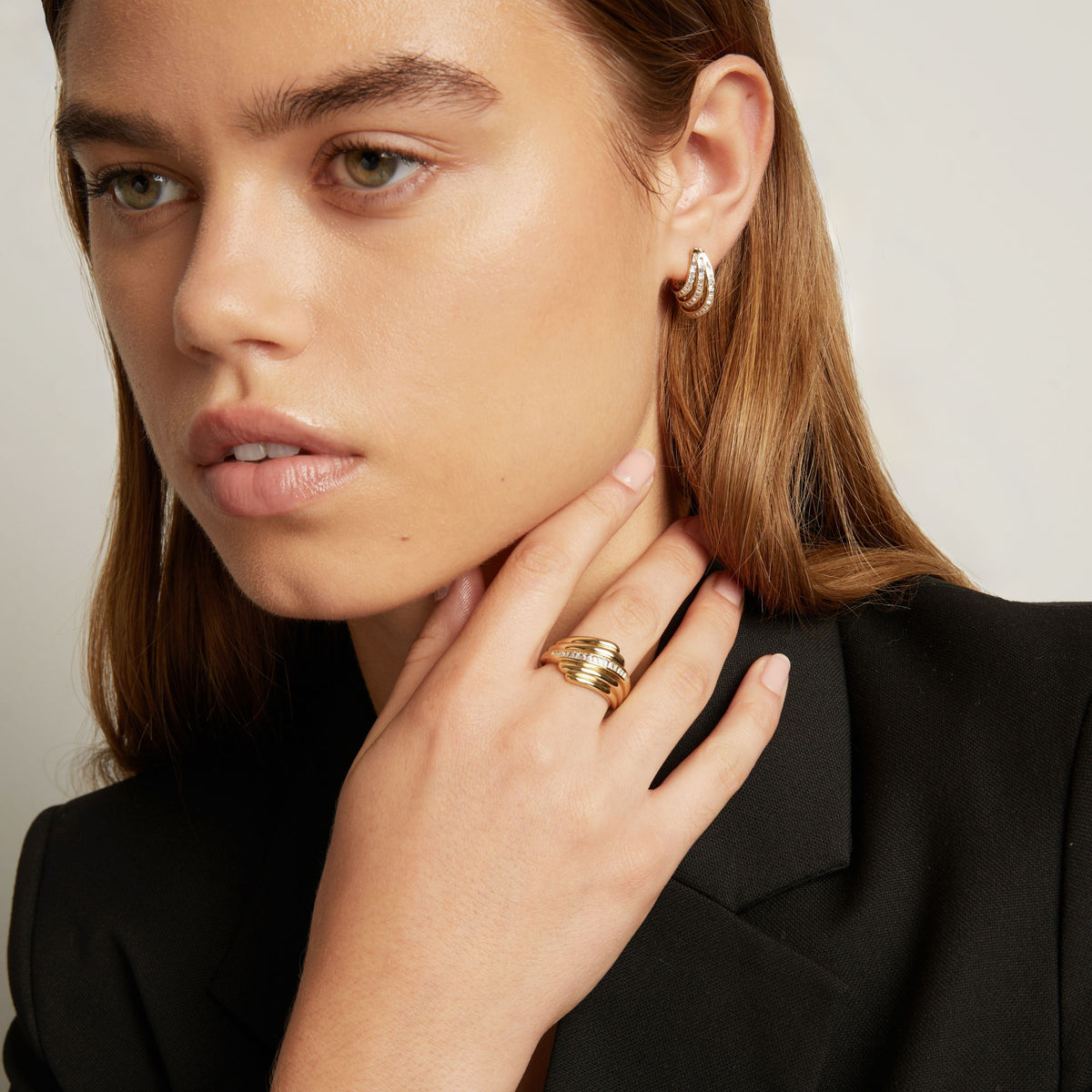 Close-up of a woman wearing a gold ring with textured horizontal bands and a baguette diamonds. Paired with gold hoop earring, a black blazer.