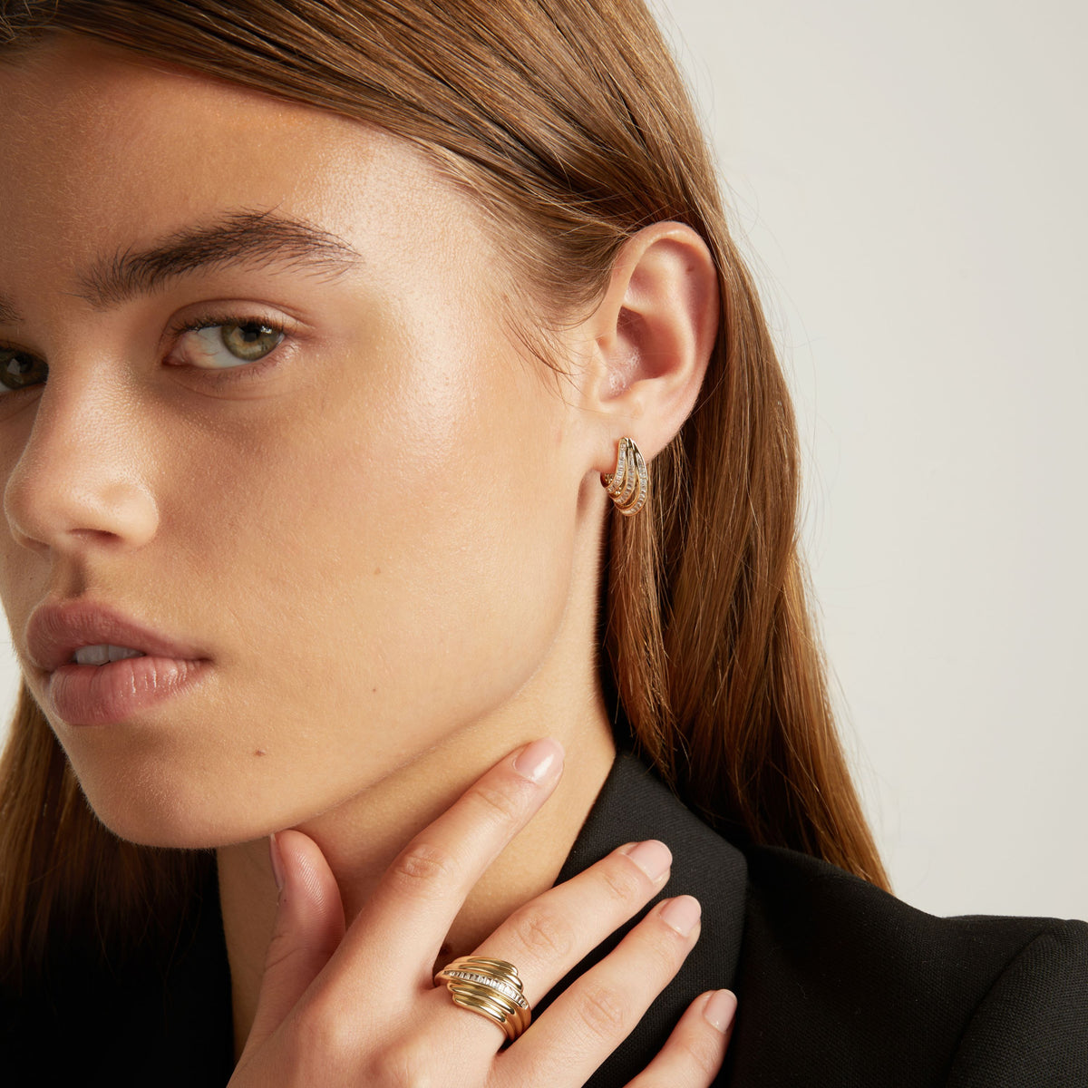 Model. A close-up of a woman with light brown hair wearing a small gold hoop earring with textured, baguette gemstone detailing, set against a plain light gray background.