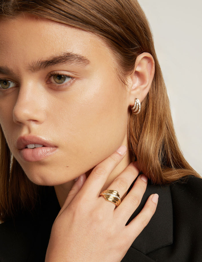 Model. A close-up of a woman with light brown hair wearing a small gold hoop earring with textured, baguette gemstone detailing, set against a plain light gray background.