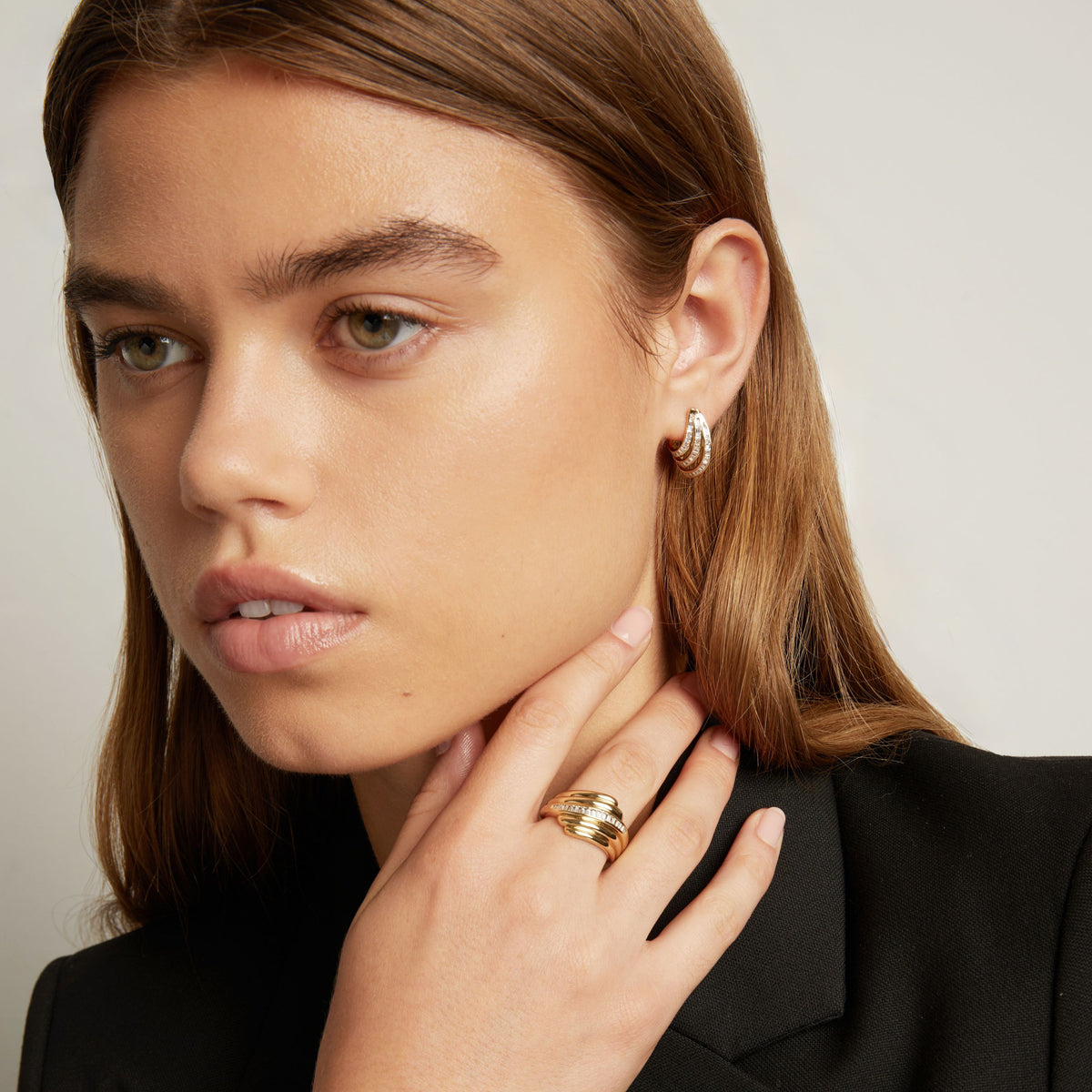Model. A close-up of a woman with light brown hair wearing a small gold hoop earring with textured, baguette gemstone detailing, set against a plain light gray background.