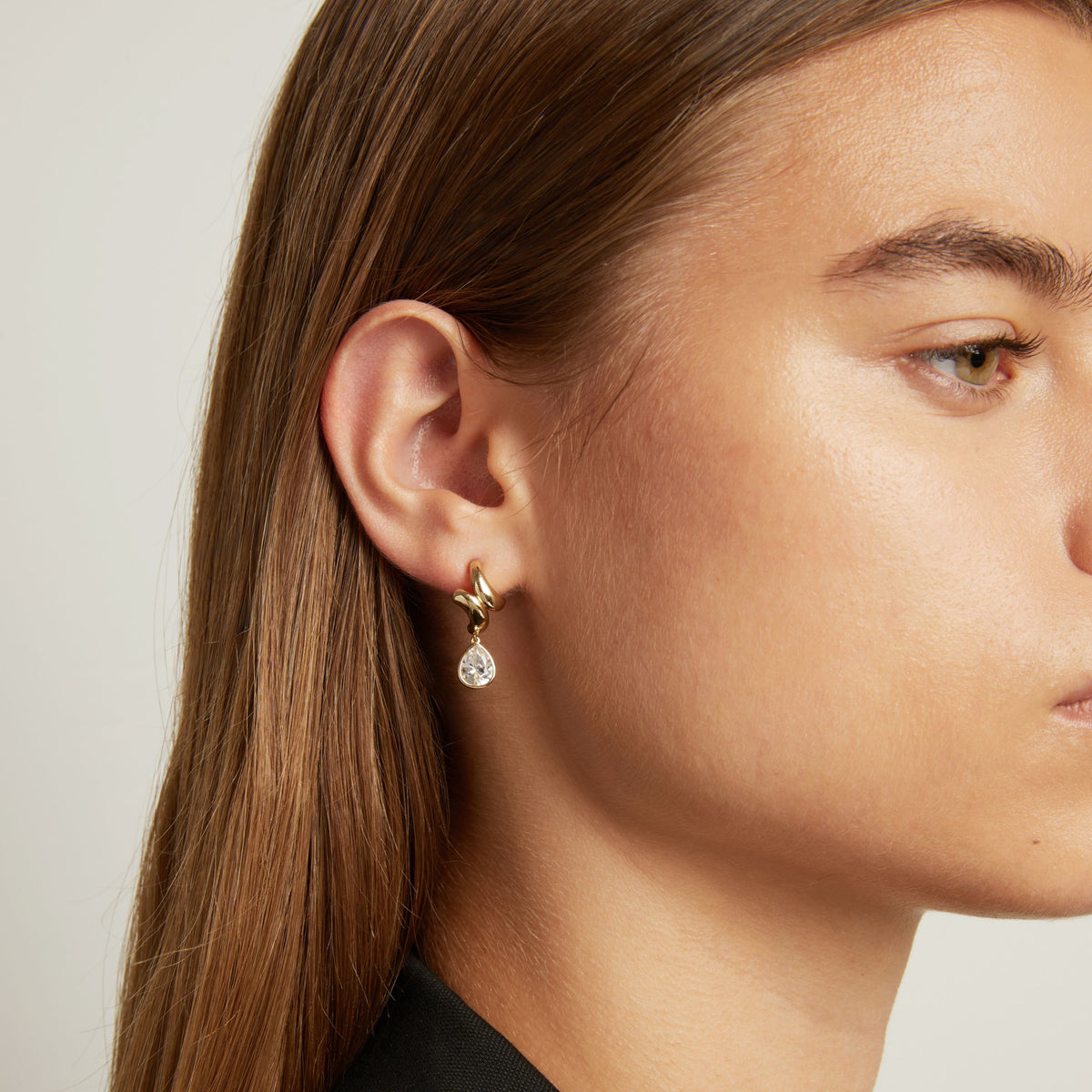 Model. A close-up of a woman with long brown hair wearing a delicate gold hoop earring with a small clear teardrop gemstone, against a light gray background.