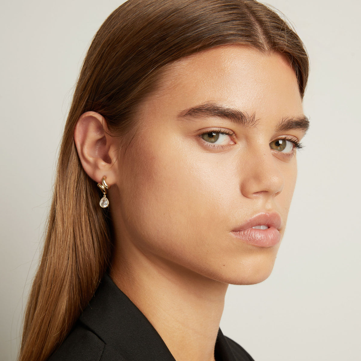 Model. A close-up of a woman with long brown hair wearing a delicate gold hoop earring with a small clear teardrop gemstone, against a light gray background.