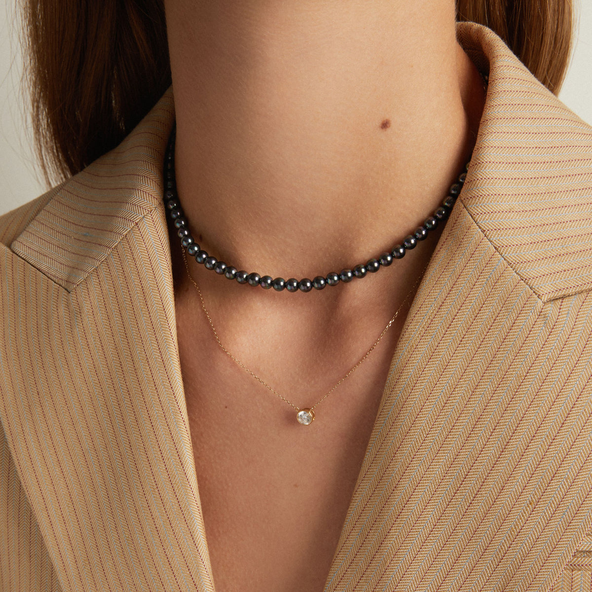 Model. A close-up of a model wearing a black pearl beaded choker necklace and a delicate gold necklace with a small clear stone pendant, styled with a beige pinstriped blazer.