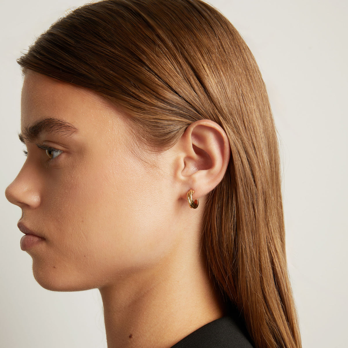 Model. Close up shot of woman with long brown hair wearing a interlocking gold hoop earring on neutral background.