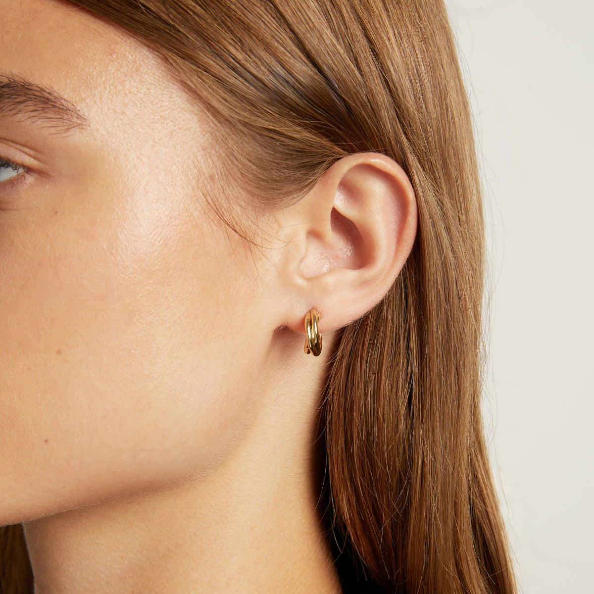 Model. Close up shot of woman with long brown hair wearing a interlocking gold hoop earring on neutral background.