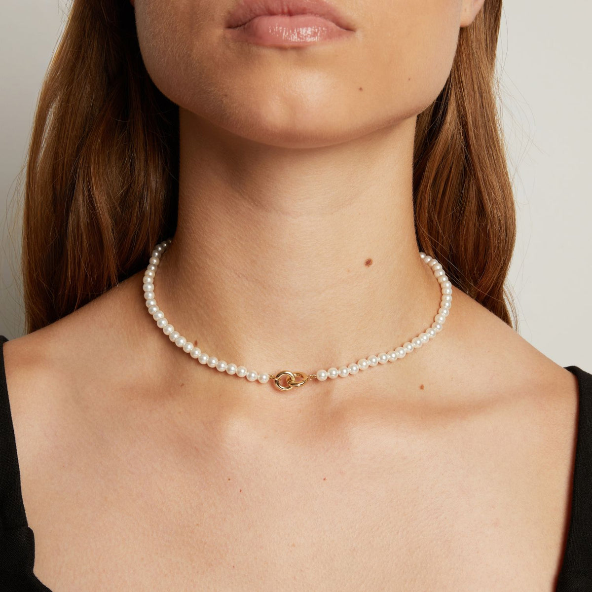 Model. A close-up of a woman wearing a delicate white pearl necklace with a small interlocking front clasp, against a neutral light gray background.