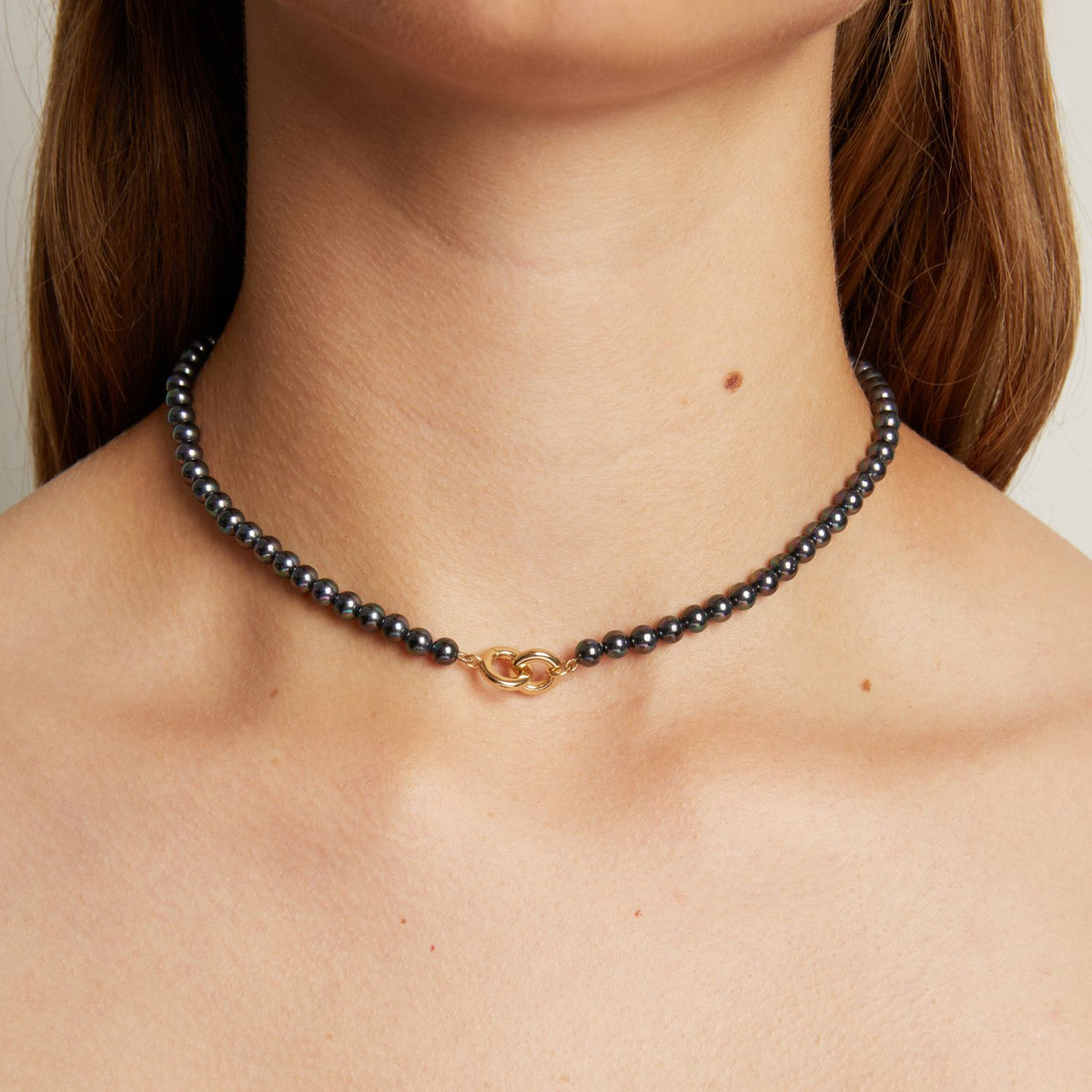 Model. A close-up of a woman wearing a delicate black pearl necklace with a small gold clasp, against a neutral light gray background.