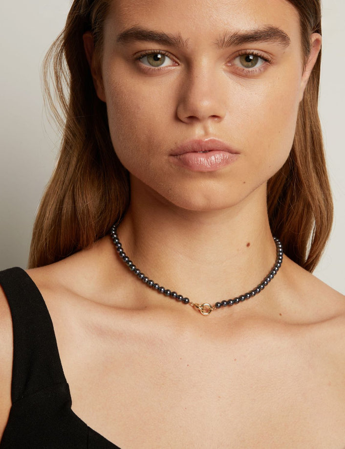 A close-up of a woman wearing a delicate black pearl necklace with a small gold clasp, against a neutral light gray background.