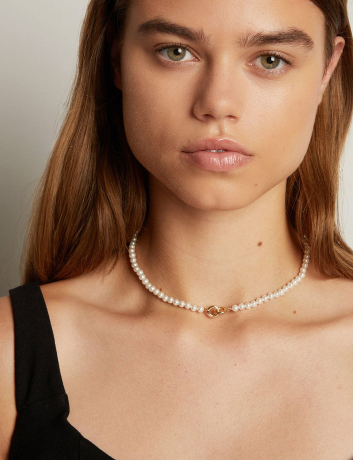 Model. A close-up of a woman wearing a delicate white pearl necklace with a small interlocking front clasp, against a neutral light gray background.