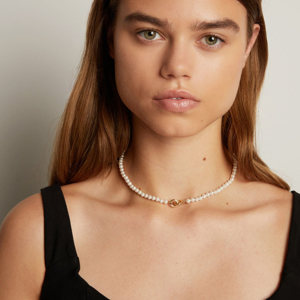 Model. A close-up of a woman wearing a delicate white pearl necklace with a small interlocking front clasp, against a neutral light gray background.