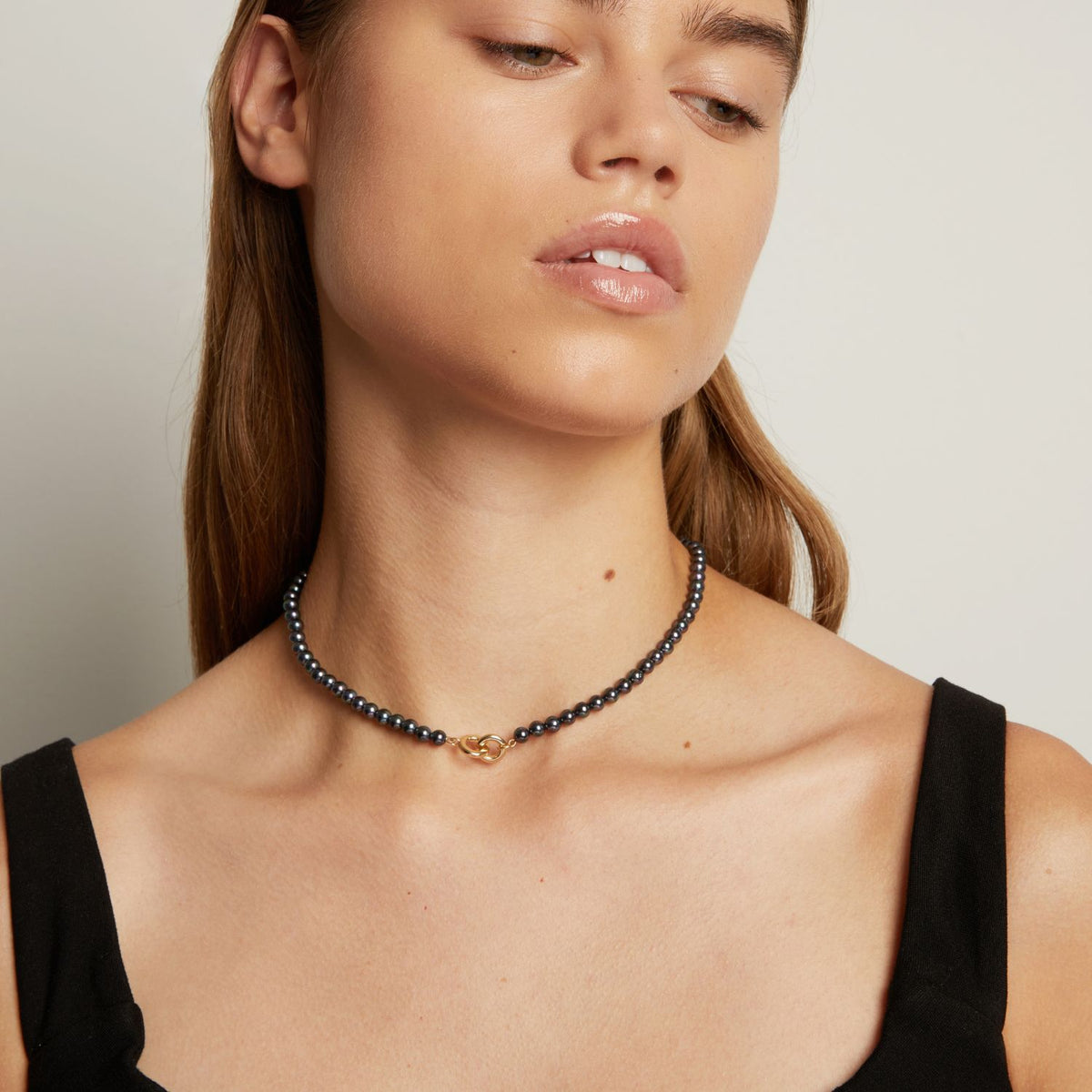 Model. A close-up of a woman wearing a delicate black pearl necklace with a small gold clasp, against a neutral light gray background.