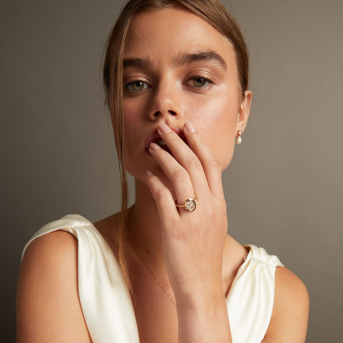 Model. A close-up lifestyle shot of a woman wearing an oval shaped diamond bezel ring on her finger, paired with a delicate gold necklace against a neutral gray background.