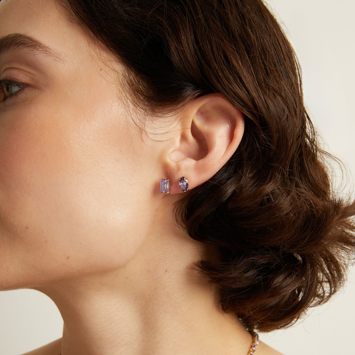 Close-up of a person wearing purple gemstone stud earrings with a neutral background. 