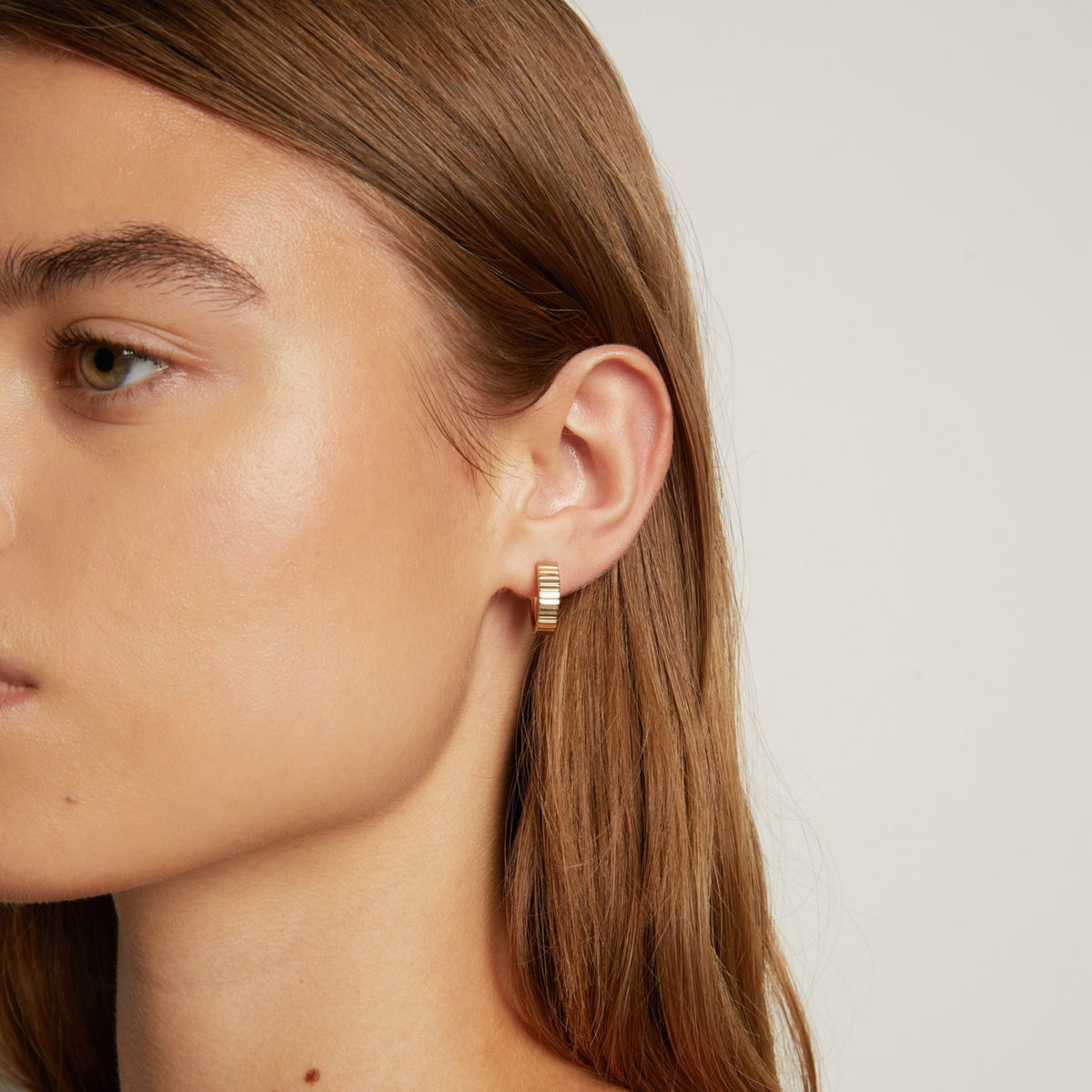Model. Close up shot of woman with long brown hair wearing a chunky gold hoop earring in neutral background.