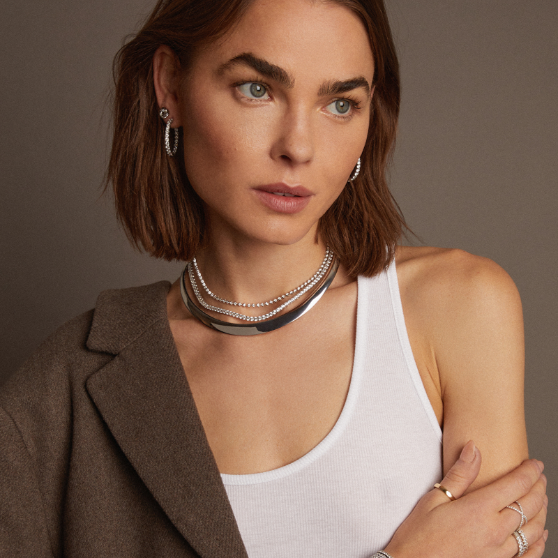Model. Woman wearing a brown blazer and white top with layered necklaces against a neutral background