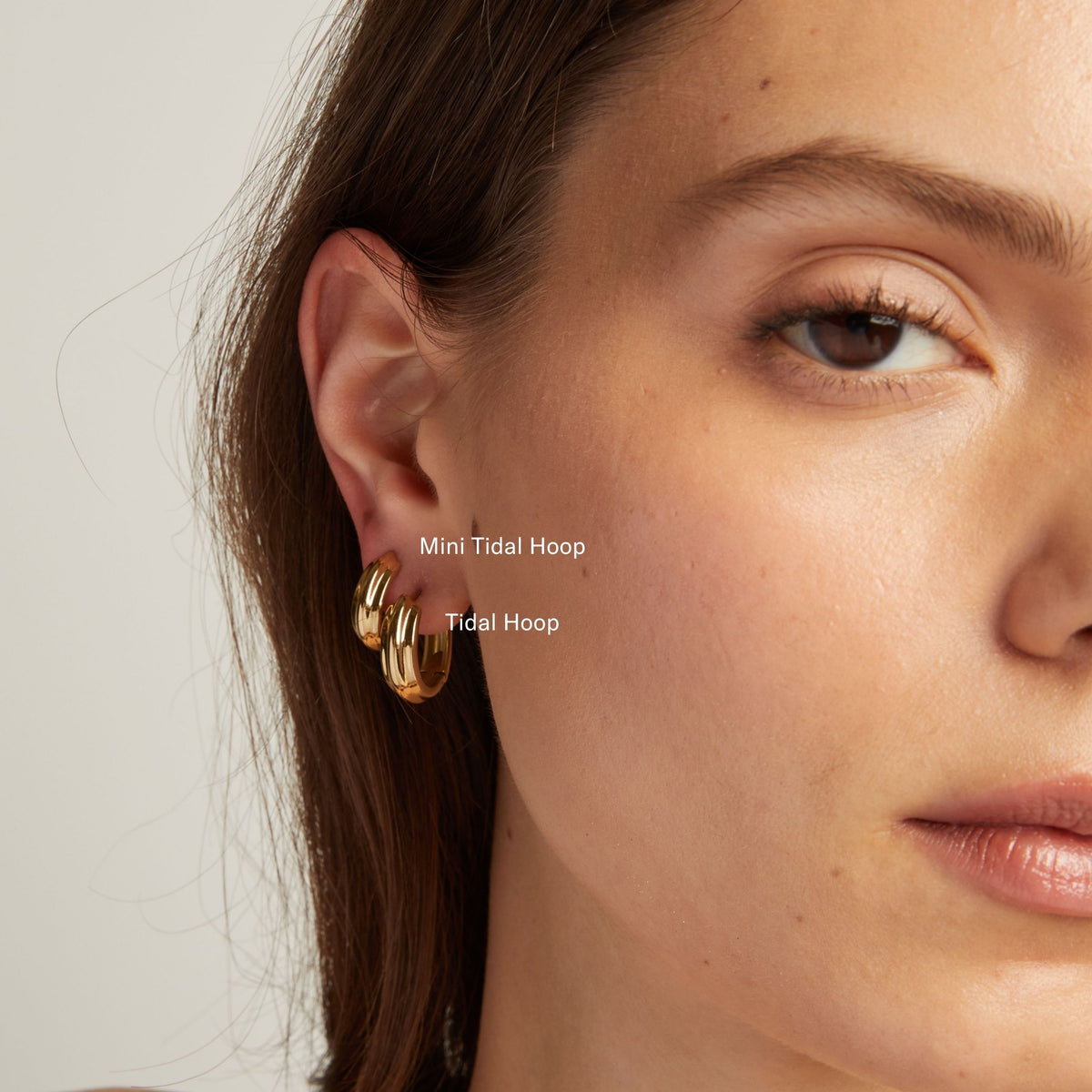 Model. Close up of a woman wearing gold hoop earrings against a neutral background