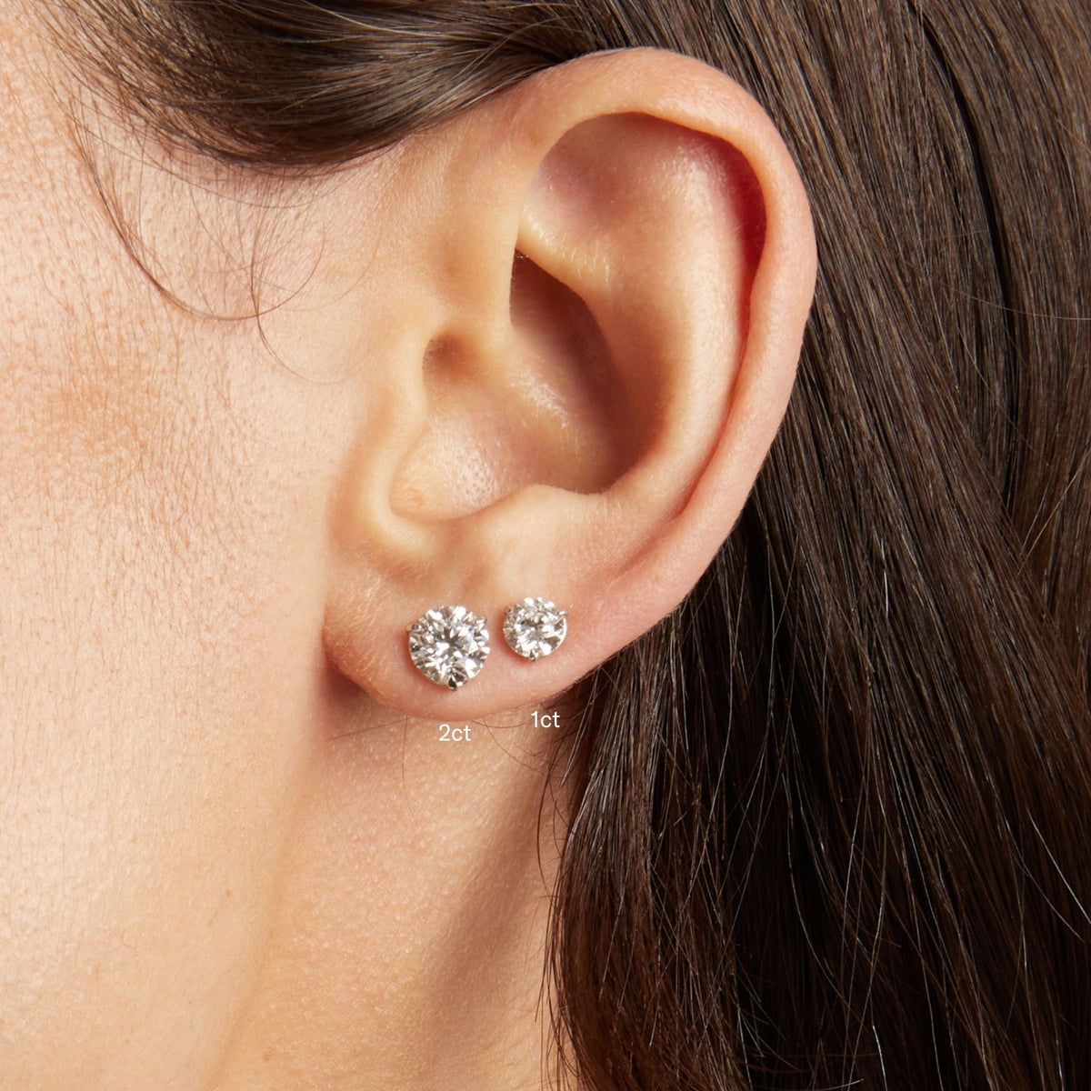 Model. Close-up of a person's ear wearing two round stud earrings, with size labels '2ct' and '1ct' near the studs.
