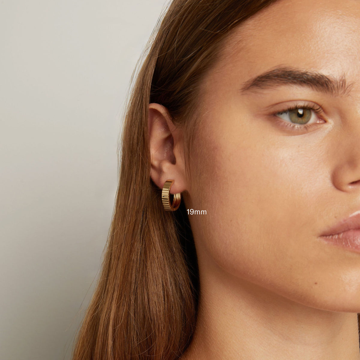 Model. Close up shot of woman with long brown hair wearing a chunky gold hoop earring in neutral background.