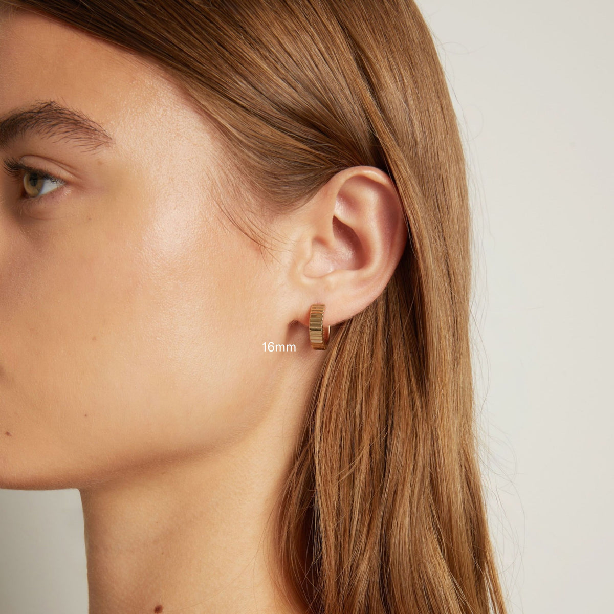 Model. Close up shot of woman with long brown hair wearing a chunky gold hoop earring in neutral background.