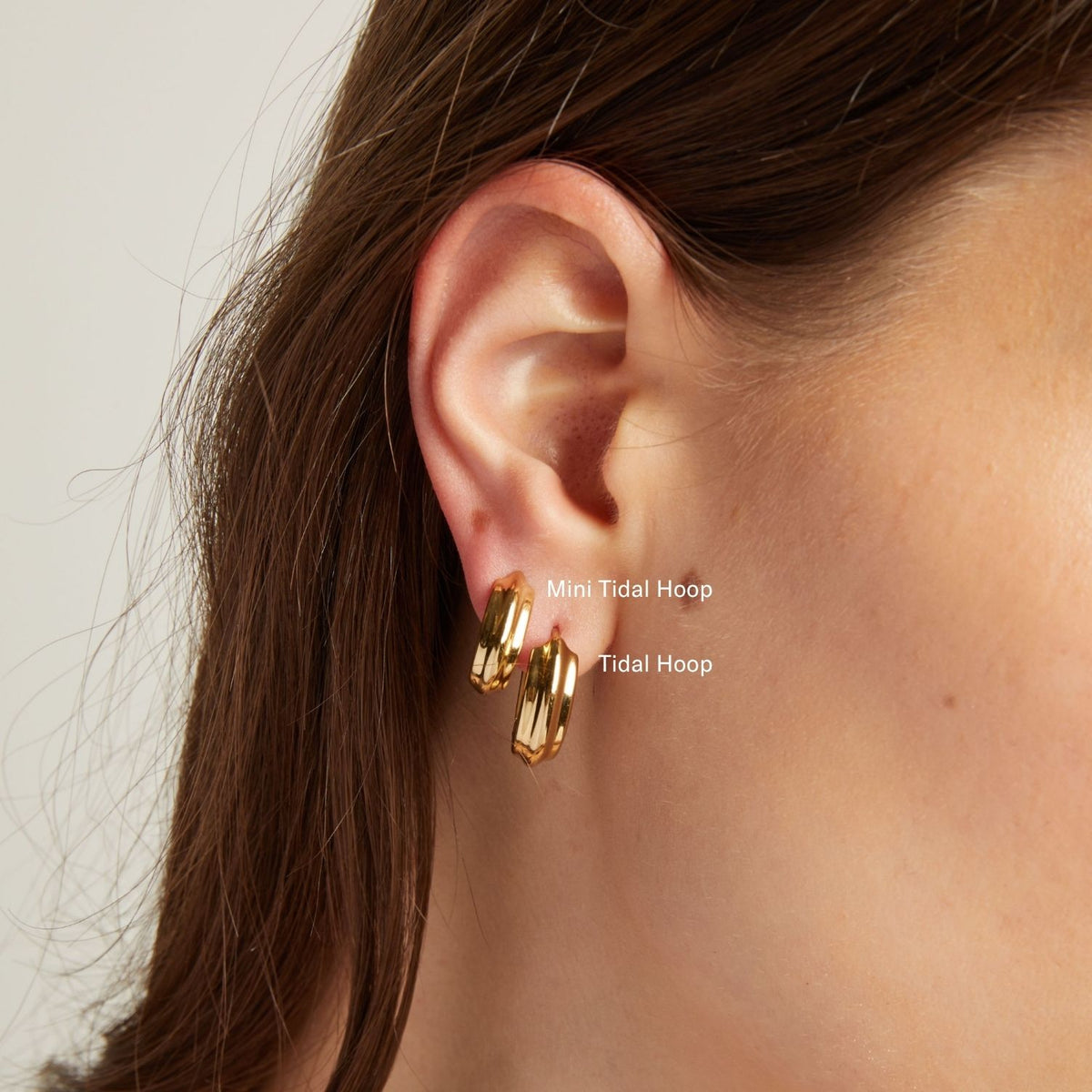 Model. Close up of a person wearing gold hoop earrings with a neutral background