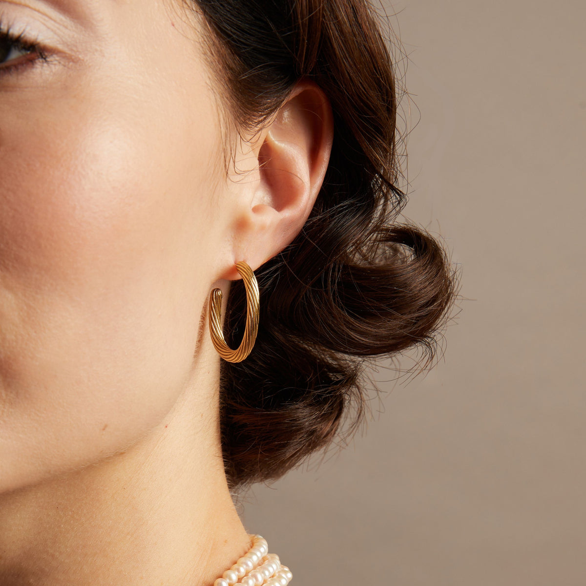 Model. Close-up of an ear wearing a gold hoop earring with a neutral background