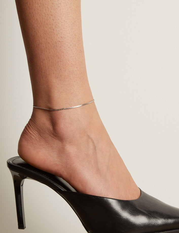 Model. Silver anklet on a foot wearing a black high heeled shoe against a neutral background.