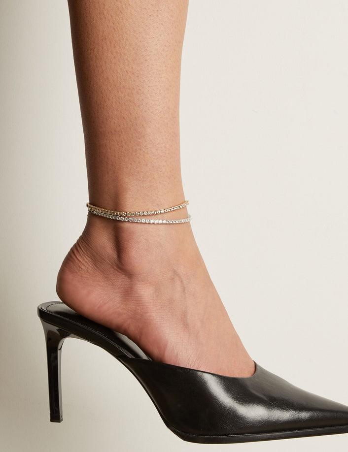 Model. Tennis anklets and black high heeled shoe worn on a foot with a neutral background.