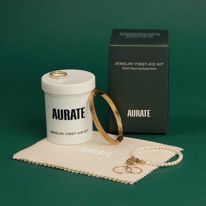 Flatlay Model A closed jar and the black packaging box of the Aurate Jewelry First-Aid Kit displayed on a green background with a branded polishing cloth, a gold bangle, a pearl bracelet, and gold rings.